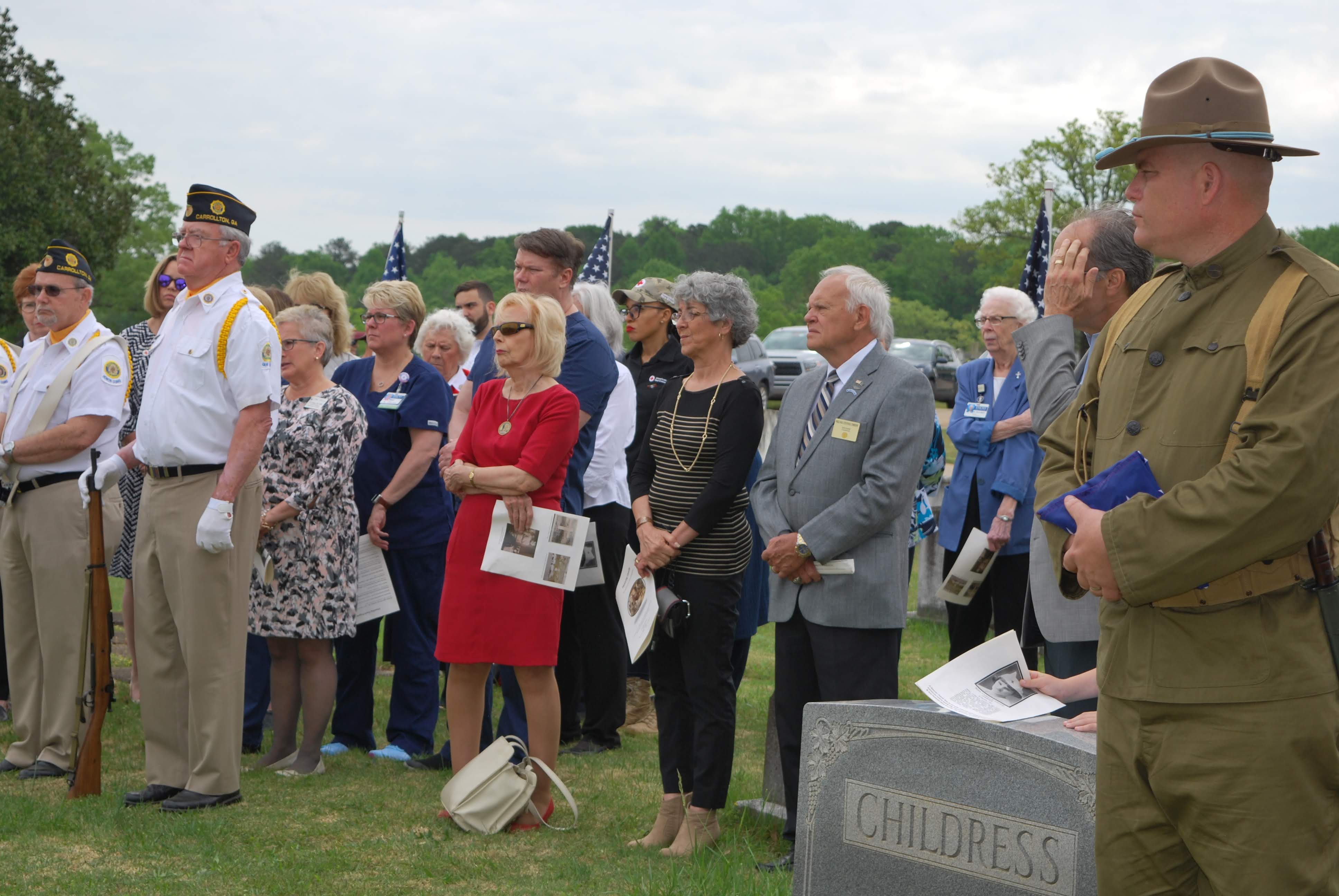 Man in authentic WW1 military uniform holds folded US flag while looking on at the ceremony. Others in attendance stand nearby.