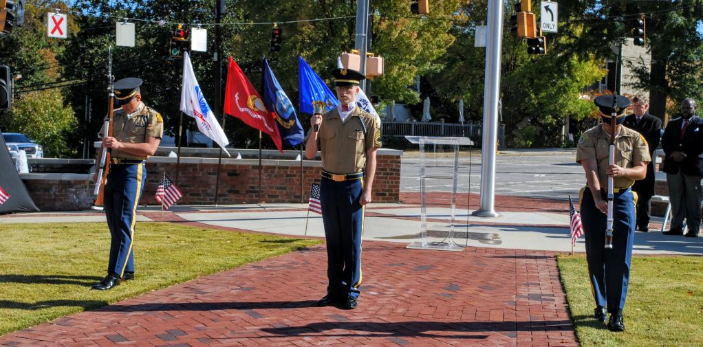 Three ROTC members perform a ceremony.  The one in the center has a bell and is ringing it.  The other two, one on each side of the bell ring, hold rifles upright in front of them and hold their heads down.
