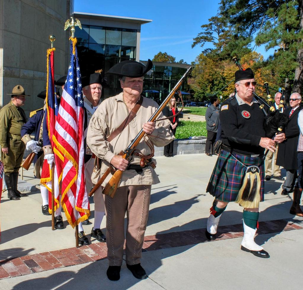 One man is dressed in revolutionary war attire and behind him are two others holding American flags.  To his left is a bagpipper