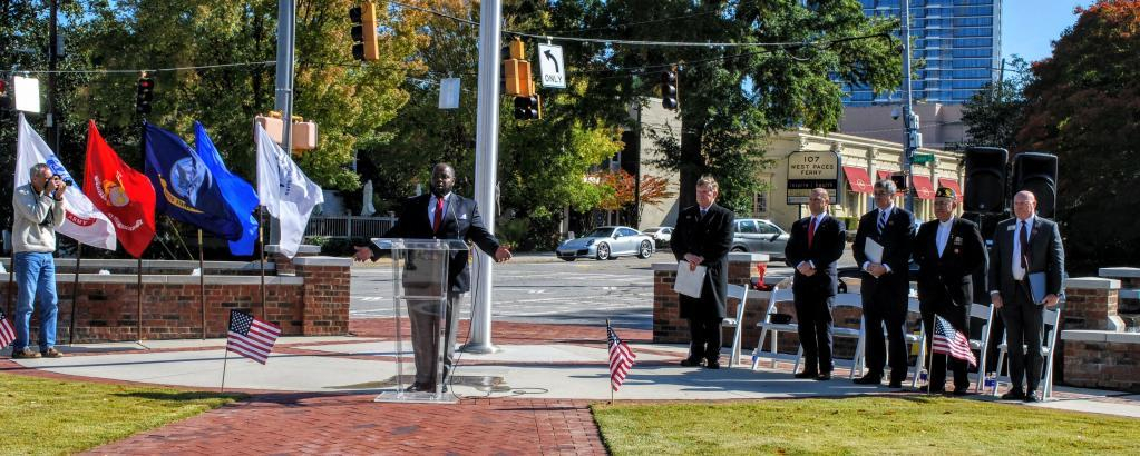 There is a speaker at a podium.  To his left are 5 mean standing in front of their chairs.  To his right are 4 flags