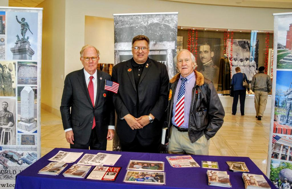 Three men stand behind a table with a purple table cloth. the table has pamphlets on it.  One man has an American flag tie and the other is holding a little American flag