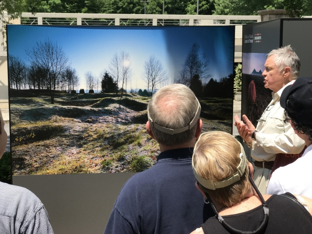 Man speaking to crowd in front of a picture of an old WW1 battlefield.  The battlefield has depressions in the ground from bombs but nature has taken over throughout the years.
