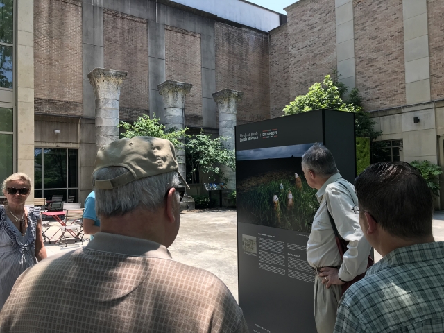 Man speaks to crowd in front of picture that looks like a color photo of soldiers walking through tall green grass