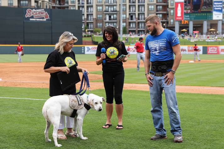 Three people with a service dog on the baseball field. Left woman holds leash of service dog, center woman has microphone and man on right looks down at service dog