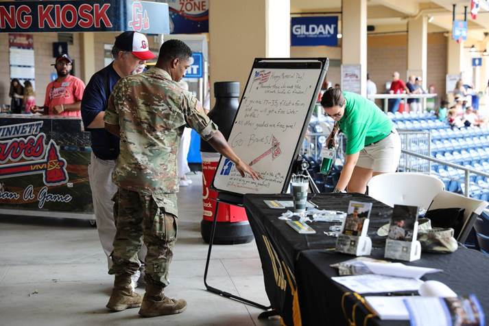 Photograph of US soldier talking with baseball fan both looking at a whiteboard on an easel that reads Happy Memorial Day