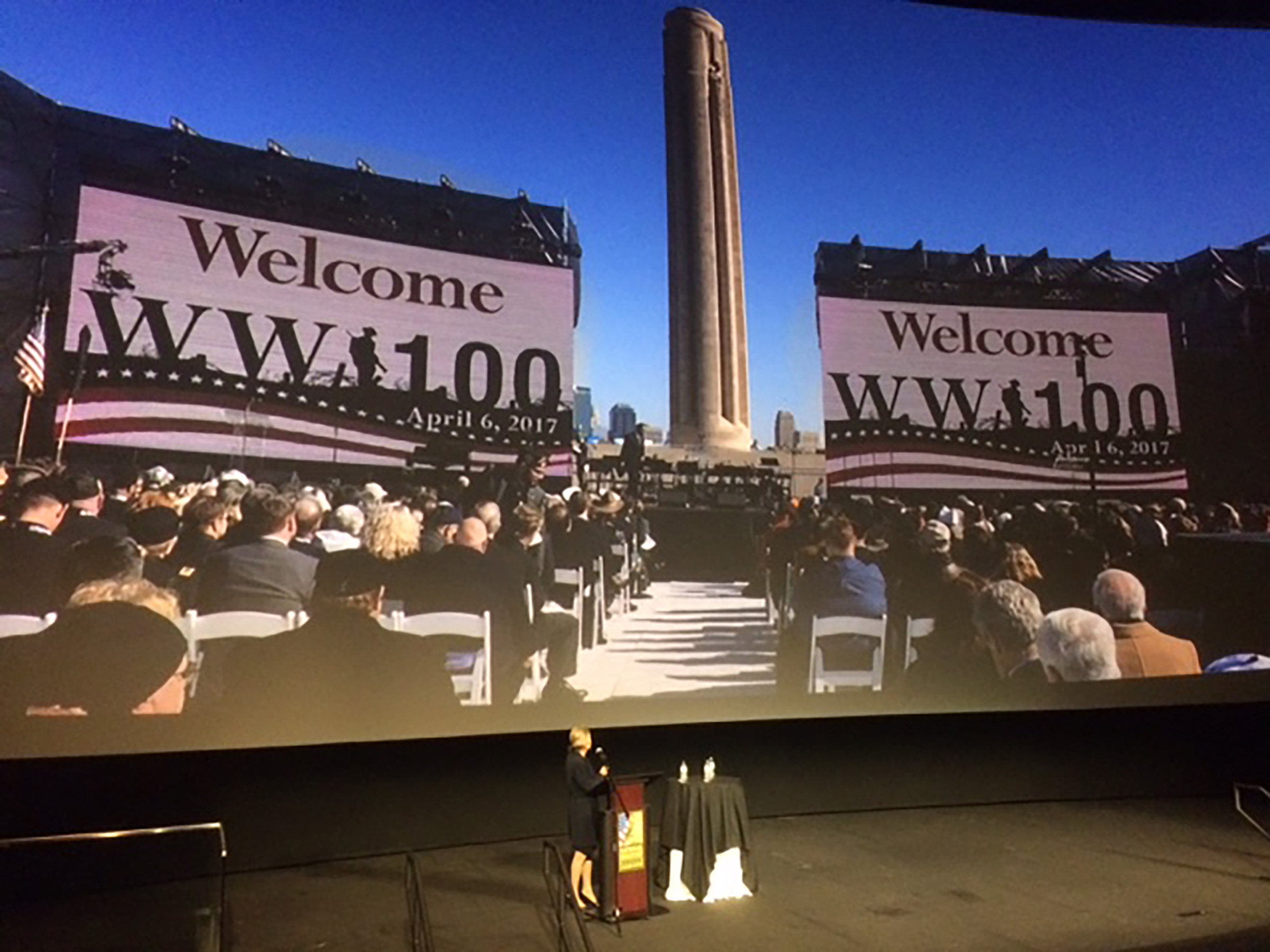 Zoomed out picture of speaker speaking on stage with projection behind